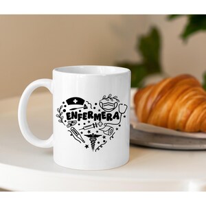 May include: White ceramic mug with a black design of a heart filled with medical symbols and the word "ENFERMERA". The mug is sitting on a white surface with a croissant on a plate in the background.