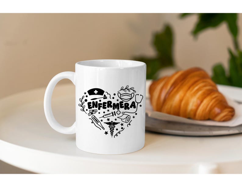 May include: White ceramic mug with a black design of a heart filled with medical symbols and the word "ENFERMERA". The mug is sitting on a white surface with a croissant on a plate in the background.