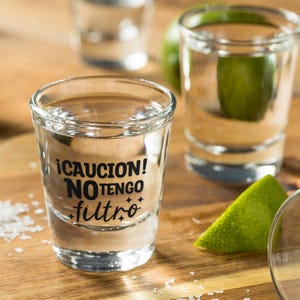 K&ouml;nnte beinhalten: Schnapsgl&auml;ser gef&uuml;llt mit einer klaren Fl&uuml;ssigkeit, wahrscheinlich Tequila, mit einer Limettenspalte und Salz auf einer Holzoberfl&auml;che. Ein Glas hat den Text "&iexcl;CAUCION! NO TENGO filtro" in Schwarz.