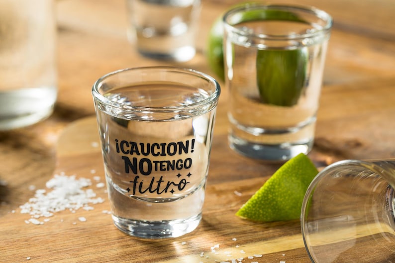 K&ouml;nnte beinhalten: Schnapsgl&auml;ser gef&uuml;llt mit einer klaren Fl&uuml;ssigkeit, wahrscheinlich Tequila, mit einer Limettenspalte und Salz auf einer Holzoberfl&auml;che. Ein Glas hat den Text "&iexcl;CAUCION! NO TENGO filtro" in Schwarz.