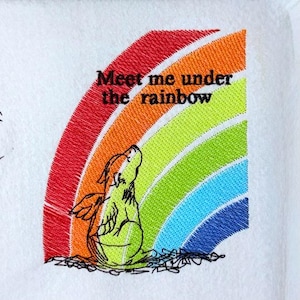 May include: Embroidered design on white fabric featuring a colorful rainbow arching over a whimsical creature with wings. The text "Meet me under the rainbow" is above the creature.