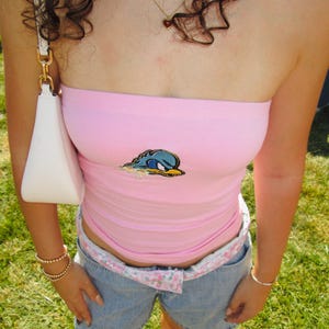 May include: A pink tube top with a cartoon graphic, paired with denim shorts and a floral belt. A white purse hangs from the shoulder. Accessories include bracelets and a necklace with a heart pendant. The outfit is worn outdoors on a grassy area.