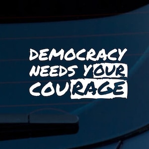 May include: White vinyl decal with the words "DEMOCRACY NEEDS YOUR COURAGE" in a bold, handwritten font. The decal is designed to be applied to a car window or similar surface, promoting a message of civic engagement.