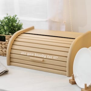 May include: A light brown wooden bread box with a roll-top lid. The text "are you bready for it?" is printed on the front. The bread box is on a white surface, with a basket of greenery and white plates in the background.