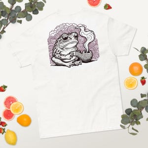 May include: White t-shirt featuring a detailed illustration of a toad smoking a pipe. The graphic is in black and white with a purple and white background. The toad is holding the pipe and smoke is rising from it.