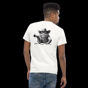 May include: A white t-shirt featuring a black and white illustration of a frog wearing a cowboy hat and holding a gun. The frog is looking to the left and the gun is pointed to the right. The frog is sitting on the ground with bullet casings scattered around it.