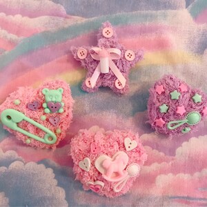May include: Four pink and purple heart-shaped felt ornaments with various embellishments. The ornaments feature a baby bottle, a teddy bear, a safety pin, and a star.