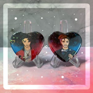 Puede incluir: Dos pines en forma de corazón con fondos coloridos e imágenes de dos personas. La persona de la izquierda lleva una chaqueta roja y la persona de la derecha lleva un traje negro.