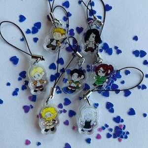 Op de afbeelding: Zeven kleurrijke telefooncharms met anime-personages. Elke charme is een transparante acrylcirkel met een zwart touwtje. De charms bevinden zich op een witte achtergrond met blauwe en roze hartjes.