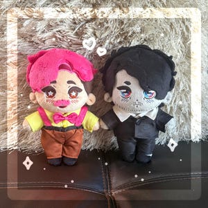 Puede incluir: Dos muñecos de peluche con rasgos faciales bordados y reflejos en forma de estrella en los ojos. El muñeco de la izquierda tiene el pelo rosa brillante, camisa amarilla, pajarita rosa y pantalones marrones. El de la derecha tiene pelo negro y traje.