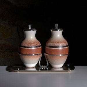 May include: Two ceramic oil and vinegar bottles with a silver metal stand. The bottles have a brown and white speckled glaze with silver bands around the middle.