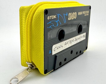 May include: A yellow zippered pouch with a black cassette tape design. The cassette tape has the text "TDK SA90 HIGH POSITION" and a handwritten label that reads "COOL AFTER DEATH 86". The zipper is silver.