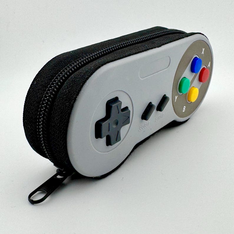 May include: A gray and black Super Nintendo controller-shaped zippered pencil case with a black zipper pull.