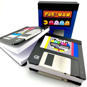 May include: A collection of retro-themed notebooks and a Pac-Man arcade-style box. The notebooks are designed to resemble floppy disks and Polaroid photos. The Pac-Man box is blue with the game's logo and characters.