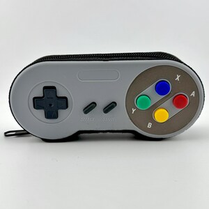 May include: A gray and black Super Nintendo controller-shaped pencil case with a zipper. The case has the classic controller buttons, including A, B, X, Y, Start, and Select.