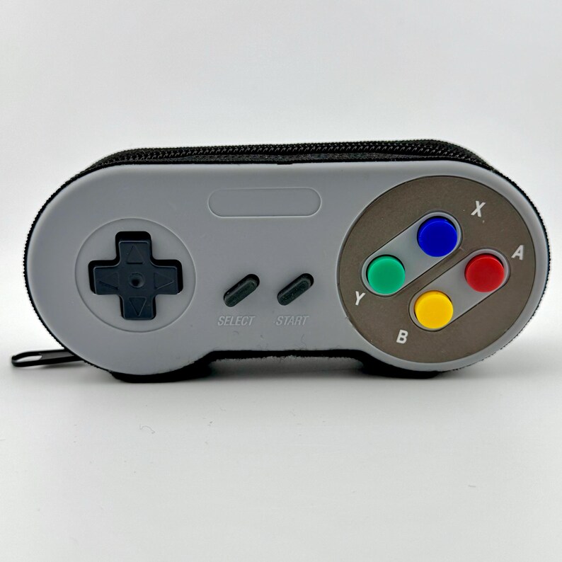 May include: A gray and black Super Nintendo controller-shaped pencil case with a zipper. The case has the classic controller buttons, including A, B, X, Y, Start, and Select.