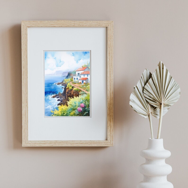 May include: Watercolor painting of a coastal scene with white buildings, red tile roofs, and a pathway along a cliff. The artwork is framed in a light wood frame and features vibrant blues, greens, and pinks. A white vase with dried palm leaves is in the foreground.