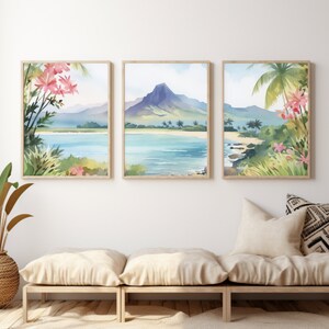 May include: A set of three framed wall art prints featuring a continuous watercolour landscape of a tropical beach, turquoise water, a prominent mountain peak, palm trees, and pink flowers, displayed in light wood frames on a white wall.