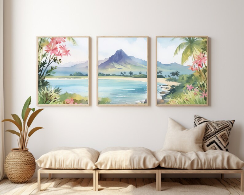 May include: A set of three framed wall art prints featuring a continuous watercolour landscape of a tropical beach, turquoise water, a prominent mountain peak, palm trees, and pink flowers, displayed in light wood frames on a white wall.