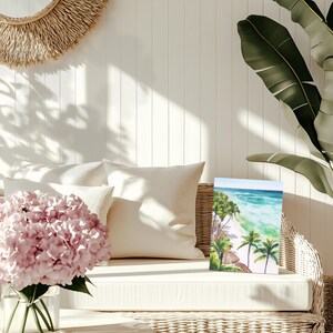 Puede incluir: Una pintura de acuarela de una escena de playa tropical con palmeras y agua turquesa. La obra de arte se exhibe en un sof&aacute; de mimbre blanco con cojines color crema. Tambi&eacute;n se ve un jarr&oacute;n de flores rosas y un tapiz de pared tejido.