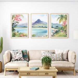 May include: A set of three framed wall art prints depicting a tropical landscape with a mountain, ocean, sandy beach, palm trees, and pink flowers. The prints are displayed in light wood frames and hung in a horizontal row on a white wall.