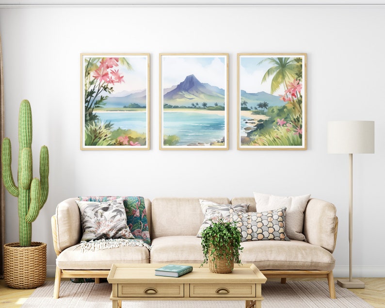 May include: A set of three framed wall art prints depicting a tropical landscape with a mountain, ocean, sandy beach, palm trees, and pink flowers. The prints are displayed in light wood frames and hung in a horizontal row on a white wall.