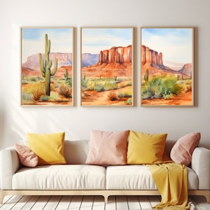May include: A triptych of framed wall art prints depicting a desert landscape with saguaro cacti and red rock mesas under a pale blue sky. The artwork features warm orange, red, and green tones, displayed in light wood frames on a plain wall.