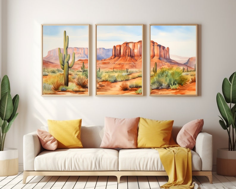 May include: A triptych of framed wall art prints depicting a desert landscape with saguaro cacti and red rock mesas under a pale blue sky. The artwork features warm orange, red, and green tones, displayed in light wood frames on a plain wall.