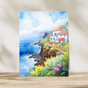 May include: Watercolor painting of a coastal scene with white buildings, red roofs, and a pathway along a cliff. The artwork depicts a vibrant blue ocean, green vegetation, and a bright blue sky with fluffy clouds. The artwork is displayed on a wooden floor.