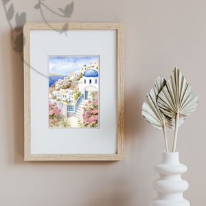 Puede incluir: Una l&aacute;mina de acuarela enmarcada que representa un pueblo costero con edificios blancos, c&uacute;pulas azules y flores de buganvilla rosas. La obra est&aacute; en un marco de madera clara con paspart&uacute; blanco, junto a un jarr&oacute;n blanco.
