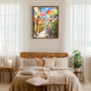 May include: A painting depicting a colourful street scene with vibrant umbrellas hanging above a cobbled street lined with buildings. The buildings are painted in shades of yellow and have green plants growing on their walls.