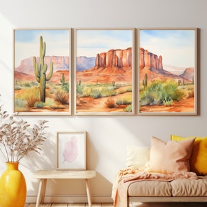 May include: A triptych of framed watercolour prints depicting a desert landscape with red rock mesas, saguaro cacti, and sparse vegetation under a pale blue sky. The artwork is displayed in light wood frames on a plain wall above a seating area.
