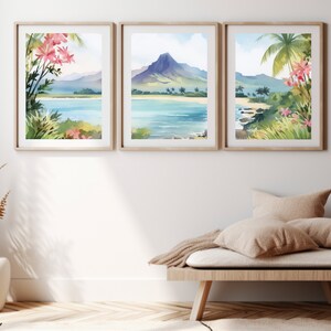 May include: A set of three framed wall art prints featuring watercolour tropical landscapes. Each print displays a coastal scene with blue water, sandy beaches, green palm trees, and pink flowers, with a central mountain peak in the middle frame.