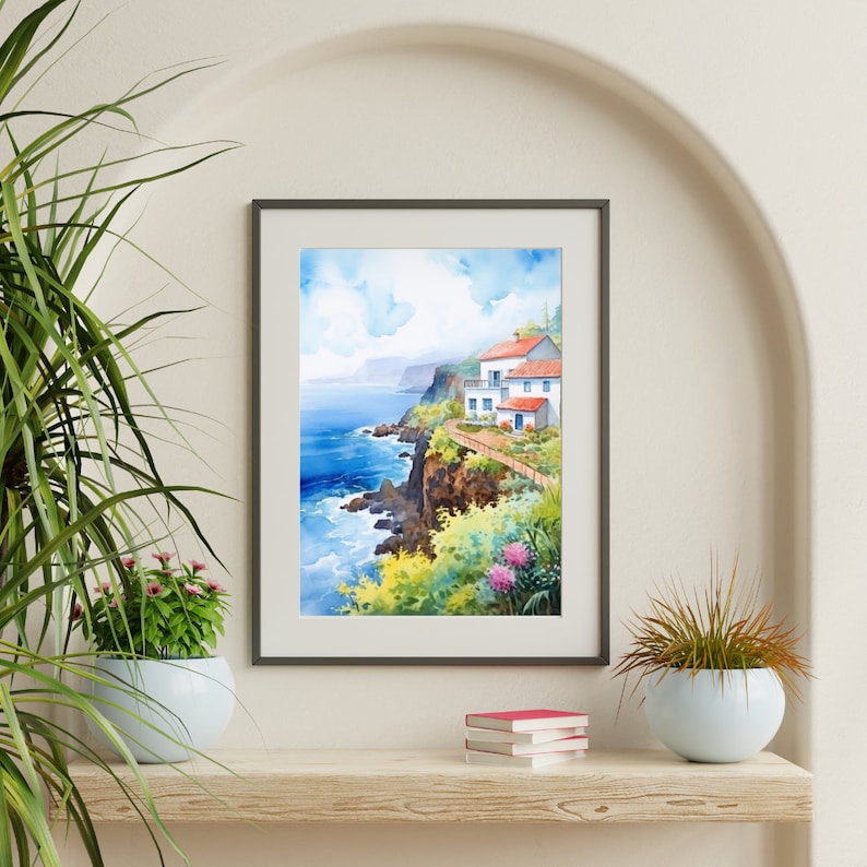 May include: Framed watercolor print of a coastal scene. The artwork depicts a white building with a red roof perched on a cliff overlooking the blue ocean. Lush greenery and colorful flowers frame the scene, with a bright blue sky above.