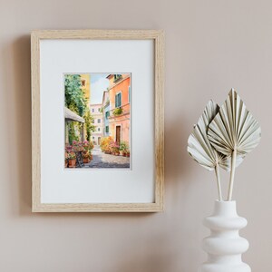 May include: A framed watercolor painting of a narrow, cobblestone street in a European city. The street is lined with colorful buildings, and there are potted plants and flowers along the sides. The painting is set against a light brown background.