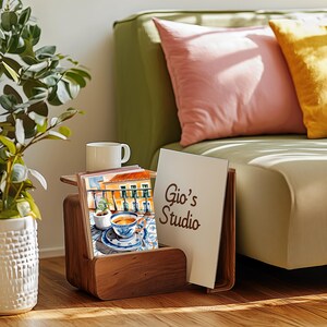 May include: A wooden magazine rack with a white card that says "Gio's Studio" and a painting of a cup of coffee on a balcony. The rack is sitting on a wooden table in front of a green sofa with pink and yellow pillows.