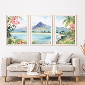 May include: A set of three framed watercolour prints depicting a tropical coastal landscape with a central mountain, blue water, sandy beaches, and pink flowers in the foreground. The artwork features light blue, green, and soft pink colour tones.