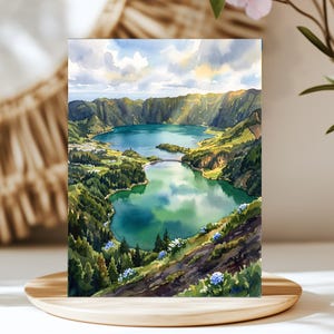 May include: A watercolor painting depicting a scenic landscape with two large lakes nestled within a deep volcanic crater. The scene features lush green hills, dense forests, and blue skies with white clouds, accented by small blue flowers in the foreground.