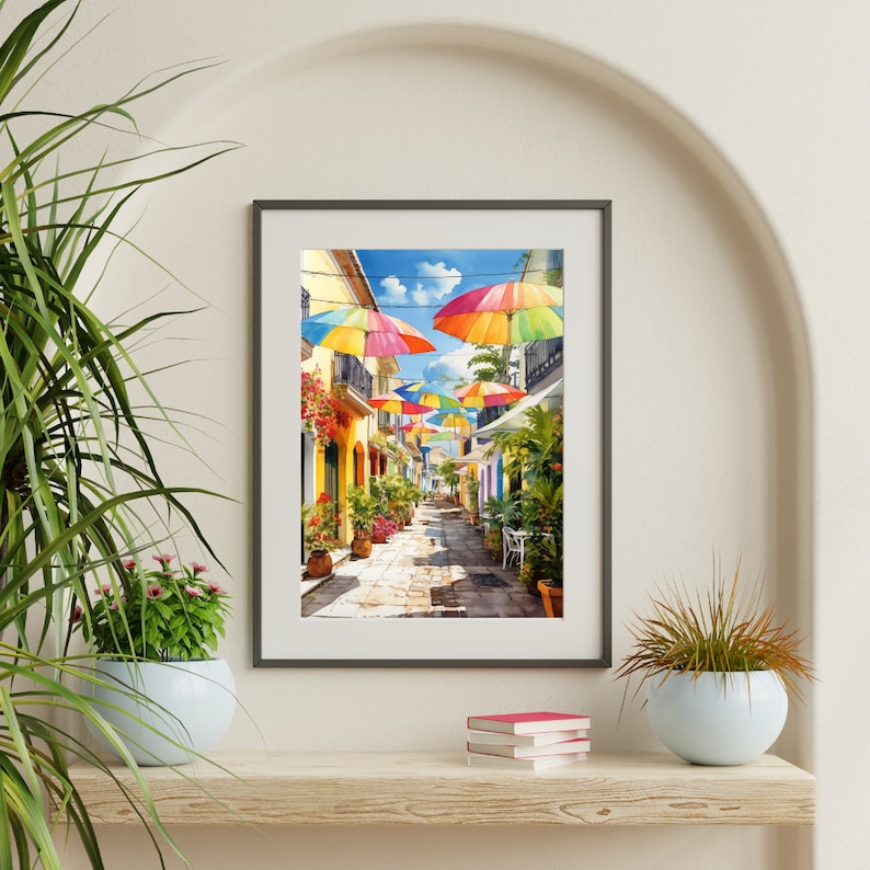 May include: A framed watercolour painting of a colourful, narrow street lined with buildings and decorated with rainbow umbrellas. The street is paved with cobblestones and has a few potted plants and flowers.