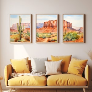 May include: A set of three framed wall art prints depicting desert landscapes with saguaro cacti and red rock formations under a light blue sky. The artwork features warm orange, red, and green tones, displayed in light wood frames on a plain wall.