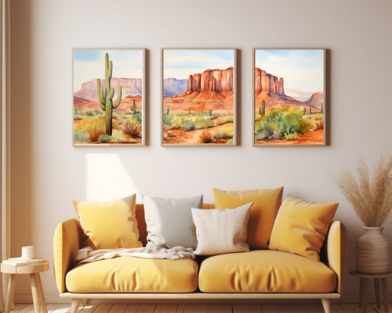 May include: A set of three framed wall art prints depicting desert landscapes with saguaro cacti and red rock formations under a light blue sky. The artwork features warm orange, red, and green tones, displayed in light wood frames on a plain wall.