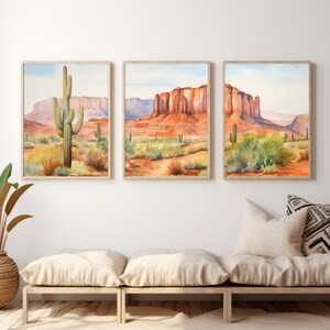 May include: A set of three framed watercolour prints depicting a desert landscape with saguaro cacti, red rock mesas, and sparse vegetation under a pale blue sky. The artwork features warm orange, red, and green tones, presented in light wood frames.