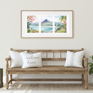 May include: A framed triptych watercolour print depicting a tropical coastal landscape with a central mountain, blue water, and lush foliage. The artwork is displayed above a wooden bench featuring two white pillows with the text welcome home.