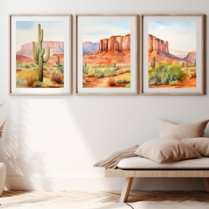 May include: A set of three framed watercolour landscape prints depicting desert scenes with saguaro cacti, red rock formations, and mesas under a pale blue sky. Each print is encased in a light wood frame, showcasing vibrant orange, red, and green tones.