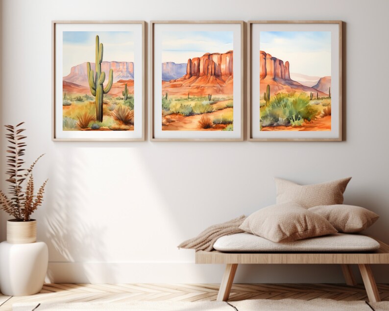 May include: A set of three framed watercolour landscape prints depicting desert scenes with saguaro cacti, red rock formations, and mesas under a pale blue sky. Each print is encased in a light wood frame, showcasing vibrant orange, red, and green tones.