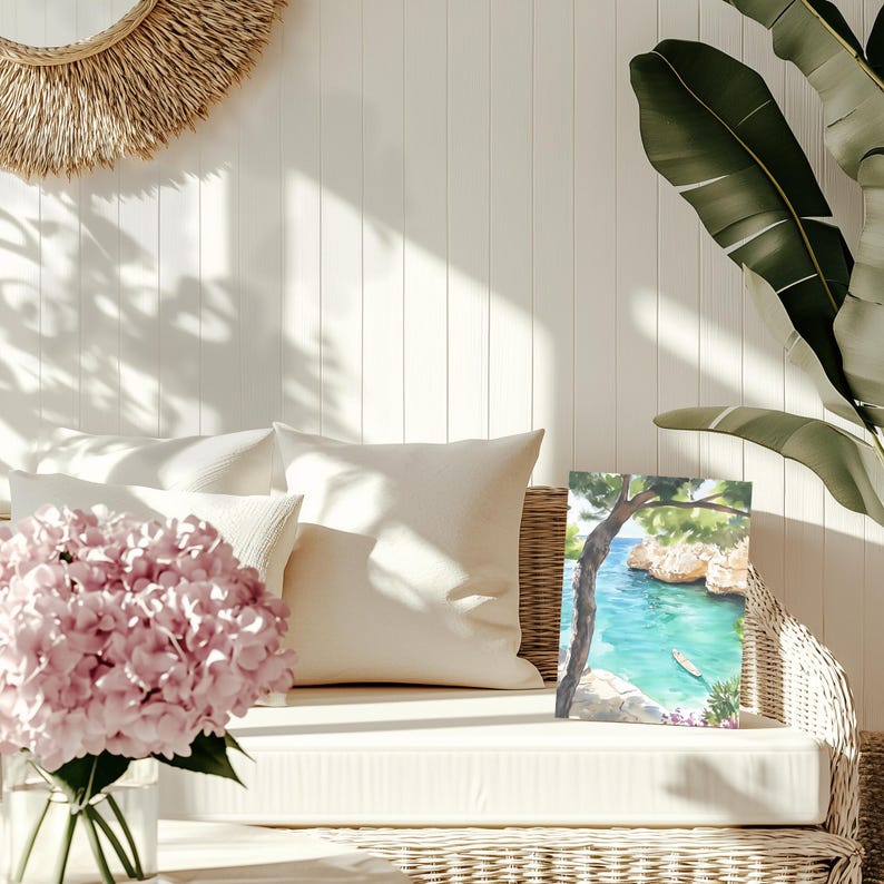May include: A watercolor painting of a coastal scene with turquoise water and a small boat. The artwork is displayed on a white wicker sofa with cream-colored pillows and a vase of pink flowers. A woven wall hanging and large green leaves are also visible.