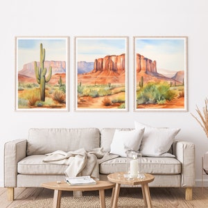 May include: A triptych of framed wall art prints featuring watercolour desert landscapes. The images depict red rock mesas, sandy terrain, and green saguaro cacti under a pale blue sky. Each print is encased in a light-coloured wooden frame.