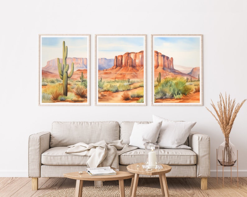 May include: A triptych of framed wall art prints featuring watercolour desert landscapes. The images depict red rock mesas, sandy terrain, and green saguaro cacti under a pale blue sky. Each print is encased in a light-coloured wooden frame.