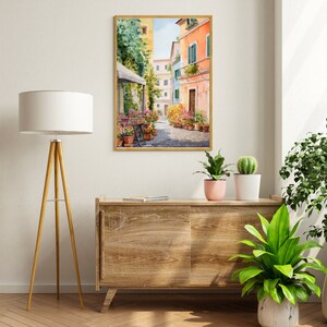 May include: A watercolor painting of a narrow street in a European city. The street is lined with colorful buildings and there are flowers in pots along the sides. The painting is framed in a gold frame and is hanging on a white wall.
