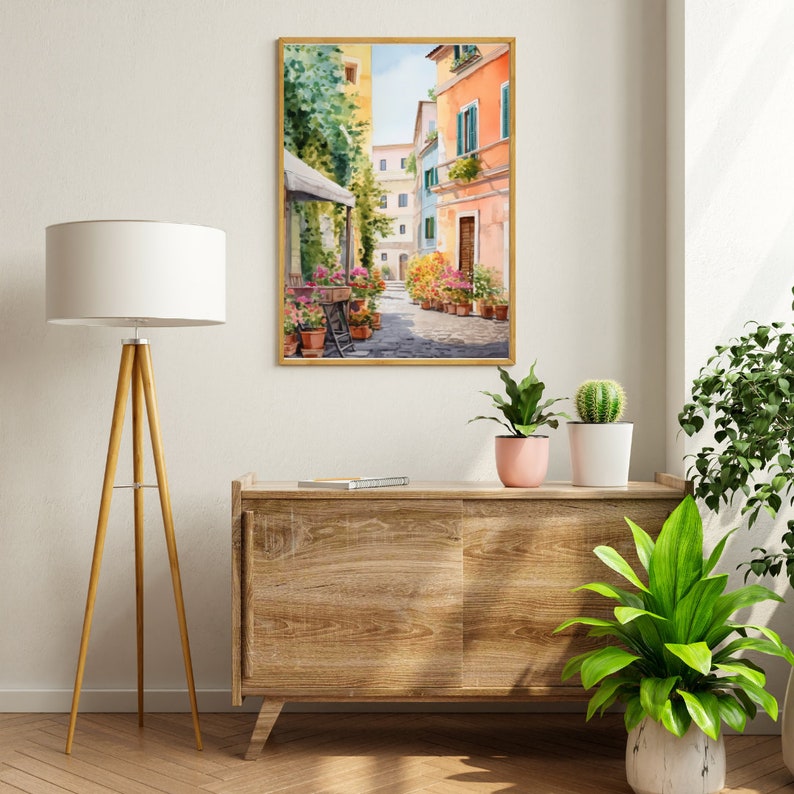 May include: A watercolor painting of a narrow street in a European city. The street is lined with colorful buildings and there are flowers in pots along the sides. The painting is framed in a gold frame and is hanging on a white wall.